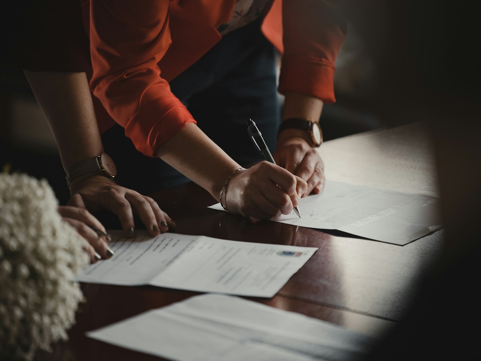 A person signing paperwork at a wooden table beside another person